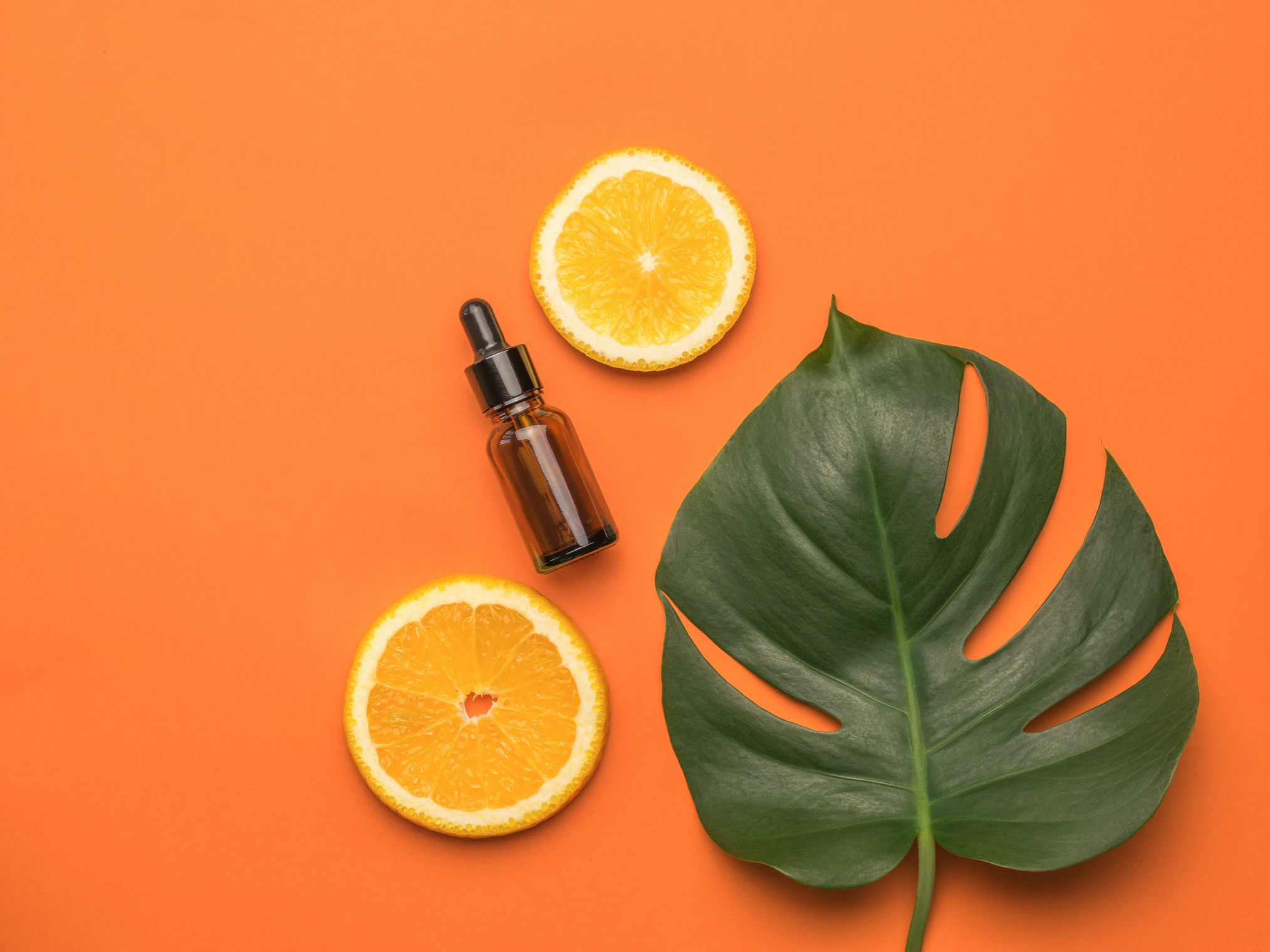Two pieces of orange, a bottle with an eyedropper and a sheet of monstera on an orange background. Flat lay.